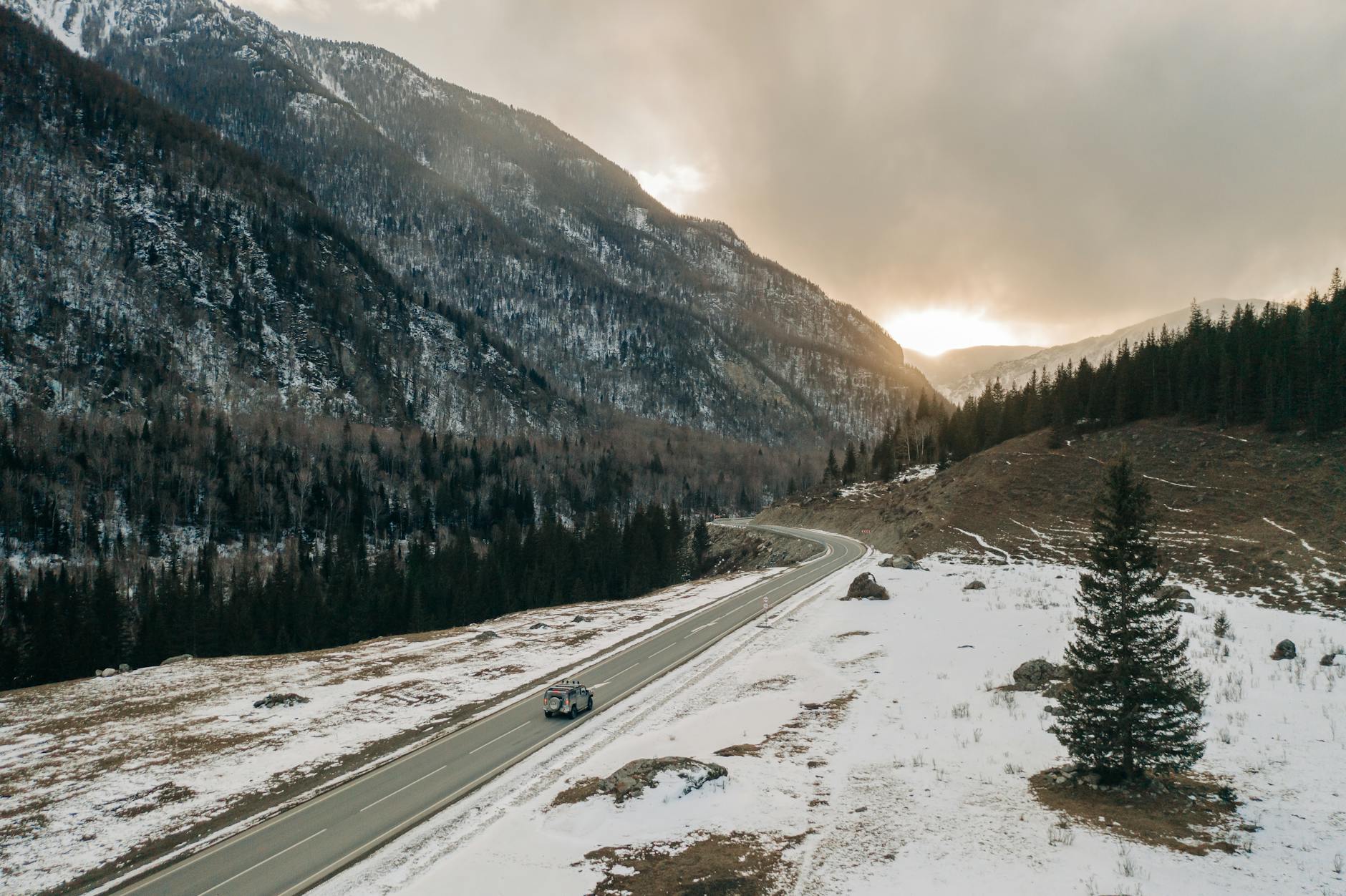 A stunning view of a road winding through snowy mountains at sunset in Russia, with pine trees and a vehicle visible.